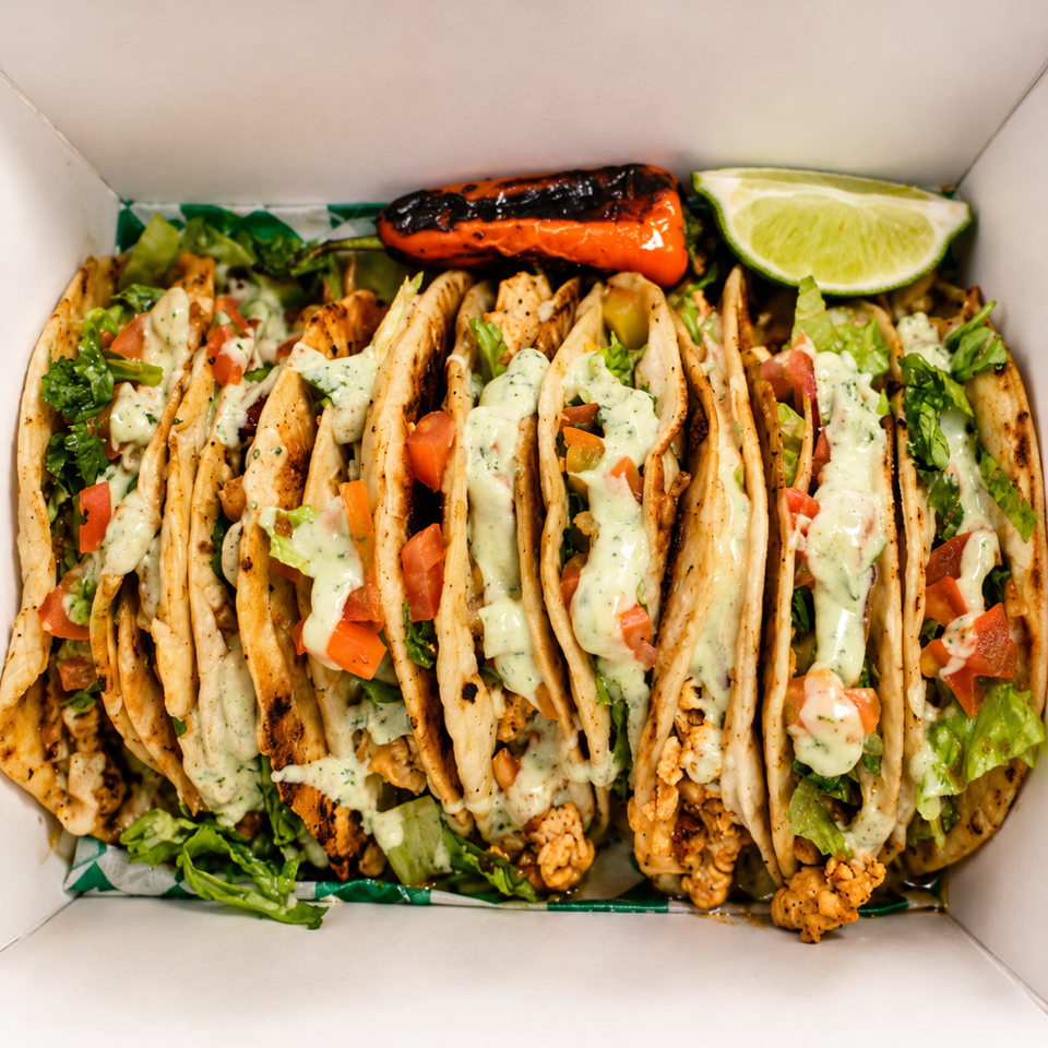 Open takeout box filled with grilled chicken tacos, lettuce, tomatoes, creamy herb sauce, roasted pepper, and lime wedge.