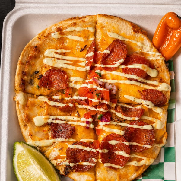 Cheesy pepperoni quesadilla topped with creamy sauce, diced tomatoes and onions, served with lime wedge and roasted peppers in a takeout box.