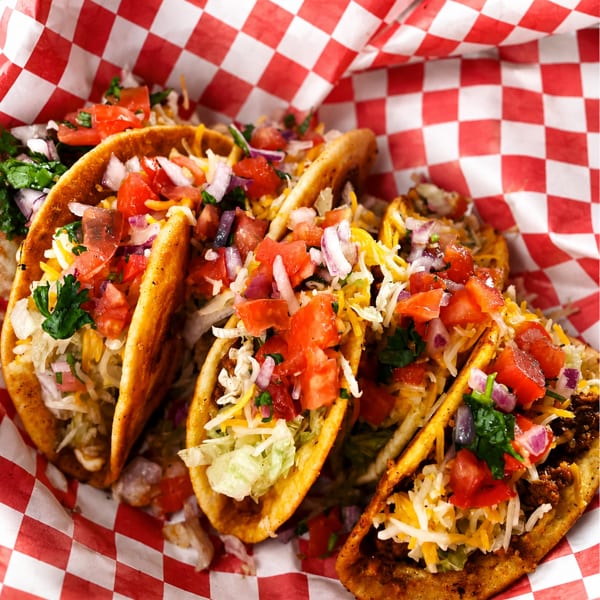 Three loaded hard-shell tacos with seasoned meat, rice, shredded cheese, tomatoes, onions, lettuce, and cilantro on checkered paper.